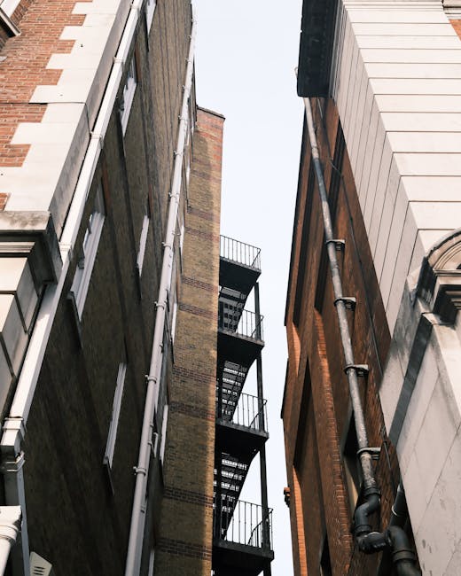 The image depicts a narrow gap between two multi-storey buildings viewed from street level, showing the vertical facades of the structures. The left building features a brick exterior with a brownish hue and several narrow windows with white frames aligned vertically. Alongside, there's a metal fire escape with black railings and diamond-patterned staircases extending from the lower to upper levels, attached to the building's facade. The right building has a white, smooth, panelled exterior with detailed stonework around a window and a black drainage pipe running vertically down the facade. The sky above is clear with natural daylight illuminating the scene, highlighting textures and subtle color variations of the brick and panelled surfaces. This setting is typical of urban environments where independent waste collection or on-site clearance might be necessary for accessing or performing rubbish removal services for buildings with limited access. The overall composition emphasizes the tight space and vertical structure common in cityscapes, aligning with the context of private waste handling and rubbish clearance solutions potentially provided by companies like Rubbish Clearance Enfield.