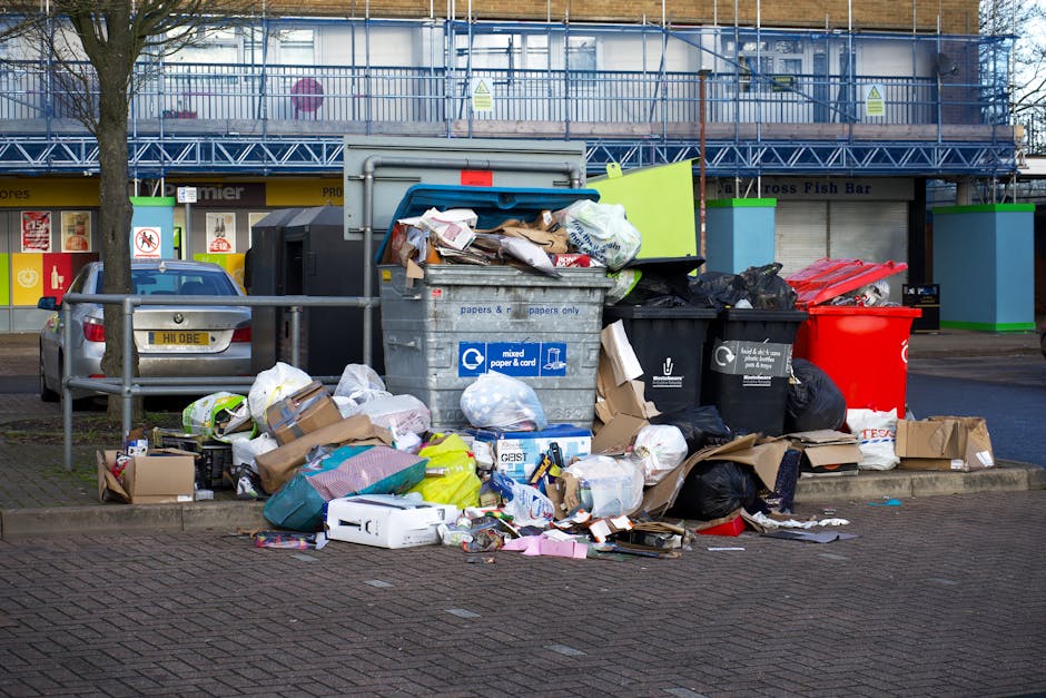 A large pile of black plastic rubbish bags, some bulging with waste, stacked against a modern tiled wall with a reflective metallic surface on the left side. The bags appear to contain household waste, with a few transparent bottles and plastic packaging visible among them. Behind the pile, there is red spray-painted graffiti reading 'XEND' on the beige tiles. The scene is set outdoors on a paved surface, with a cloudy sky partially visible in the background. The image reflects a situation where independent waste collection or private rubbish removal services, such as those provided by Rubbish Clearance Enfield, might be required to manage and clear waste piling up in an urban environment, especially around access points for flats above shops or commercial premises.