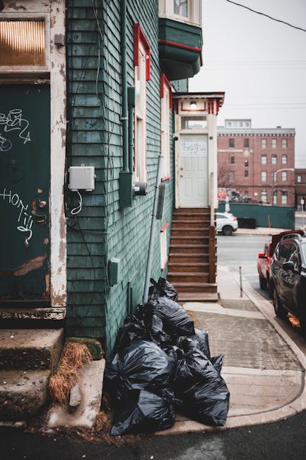 In the foreground of the image, there are several tightly packed black garbage bags piled against the side of a building with teal weatherboard siding, which exhibits a textured, slightly weathered appearance. The bags are stored at the base of a small set of wooden stairs leading up to a white door with decorative trim, positioned on the front of the building. To the left, part of a dark, graffiti-covered door with peeling paint and rusted hinges is visible, indicating an older, urban environment. Around the bags, there is a small concrete sidewalk and a patch of ground layered with ice or snow. The building features a small window with a white frame, and electrical meters or utility boxes are mounted on the exterior wall, hinting at private waste handling or independent rubbish collection services. In the background, a parking lot with a few parked cars, including a dark-colored vehicle, and a large brick building, suggest an industrial or residential area across a street. The overall scene portrays a typical urban setting where rubbish is temporarily collected outside properties, supporting alternative waste management practices outside of municipal collection, with natural light providing a neutral, daytime atmosphere. Rubbish Clearance Enfield’s service focus on rubbish removal is linked to this type of outdoor waste storage and clearing.