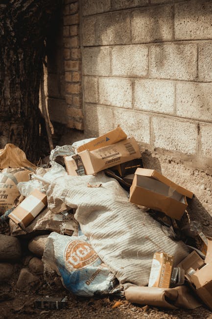 A pile of discarded cardboard boxes, some flattened and others partially intact, is seen resting against a weathered brick wall and a concrete block wall, adjacent to a tree trunk. The boxes vary in size and are light brown with visible printed labels and barcodes. Some boxes are torn or crumpled, creating an uneven surface intermingled with crumpled paper and plastic packaging. The ground beneath the rubbish consists of dirt and small stones, with a suggestion of additional debris scattered nearby. The scene appears to be an outdoor area, possibly part of a private property or service site where waste has been temporarily accumulated. The lighting is natural, highlighting the textures of the cardboard, the roughness of the brick wall, and the surrounding environment. This image exemplifies a household or commercial waste collection point, commonly handled through independent rubbish removal services like Rubbish Clearance Enfield, aligning with the theme of alternative waste management or on-site clearance outside local authority collection zones.