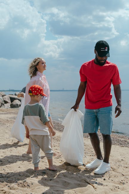 A man and two children gathering litter on a sandy beach near a body of water under a partly cloudy sky. The man, positioned on the right, wears a black cap, red T-shirt, denim shorts, and white sneakers, holding a white rubbish bag in one hand. The children, a young girl with curly blonde hair in a light jacket and beige trousers, and a young boy with a red cap, light blue T-shirt, and beige trousers, are standing nearby, appearing to assist in collecting waste. The background features a rocky shoreline with large stones and a calm water expanse extending to the horizon, with some distant land visible. The scene is lit by natural daylight, emphasizing the neutral tones of the sand and water, creating a quiet, outdoor environment suitable for community cleanup activities, similar to the efforts of local waste removal services like Rubbish Clearance Enfield for alternative disposal solutions.