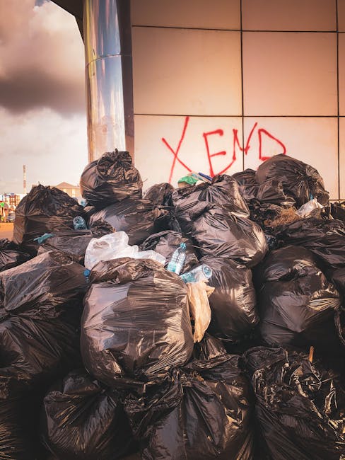 A large pile of black rubbish bags stacked against a tiled exterior wall, with some bottles and waste visible around and on the bags. The background shows part of a building with reflective metal panels and graffiti with the word 'XEND' spray-painted in red. The scene appears to be an outdoor urban environment, possibly on a pavement or driveway, with natural lighting casting shadows and highlights on the bags and wall. The image reflects an example of waste awaiting collection, illustrating private or independent rubbish disposal efforts that Rubbish Clearance Enfield offers as an alternative to local authority rubbish removal services.
