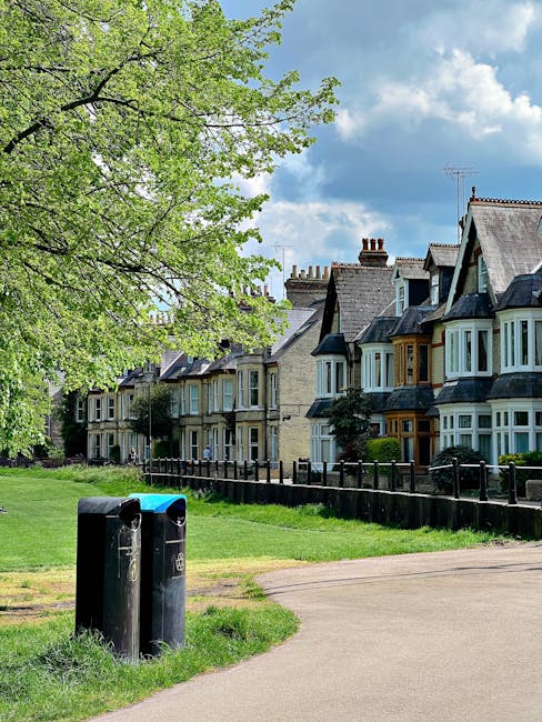 A row of traditional terraced houses with brick facades, bay windows, and slate roofs are visible in the background, with some chimneys protruding from the rooftops. In the foreground, a green grassy area bordered by a black metal fence separates the residential row from a paved footpath. To the left, a large tree with lush, light green leaves extends partly across the frame, providing partial shade. Near the base of the tree, two black rubbish bins with blue lids are positioned close together on the grass, indicating a nearby waste collection point. The sky overhead is partly cloudy, with patches of blue sky peeking through the white and gray clouds, creating a bright yet slightly overcast atmosphere characteristic of a typical day in a suburban area. The scene suggests a quiet neighborhood where waste disposal may involve private on-site clearance services such as those offered by Rubbish Clearance Enfield, aligning with alternative waste handling methods away from municipal collection, supported by the visible bins and residential setting.