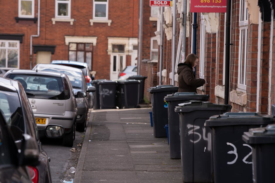 A suburban street scene featuring a row of black wheelie bins placed along the pavement adjacent to residential brick houses. The bins are lined up closely, with some visibly labeled with white spray paint numbers. The driveway in the foreground shows parked cars, including a small silver hatchback and a slightly larger vehicle nearby, with reflections visible on their surfaces. In the background, a woman dressed in a dark coat is seen standing beside one of the bins, possibly managing waste disposal or rubbish collection. The brick facades of the houses display a traditional Victorian style, with some windows and exposed rockery on the building fronts. The environment is outdoors, with natural daylight illuminating the scene, suggesting daytime hours. This scene visually relates to private waste handling services often provided by rubbish clearance companies such as Rubbish Clearance Enfield, supporting the process of alternative rubbish removal outside of local authority collection. The image emphasizes the typical setting for on-site rubbish clearance involving external waste storage and collection preparation.
